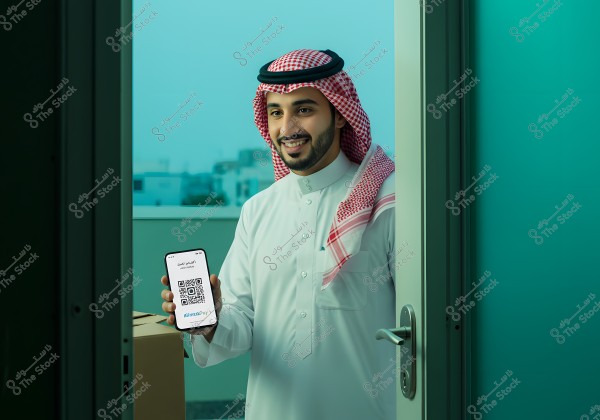 An image of a man wearing a white traditional garment and a red and white headscarf, opening a door and smiling. He is holding a smartphone displaying a digital payment app with a QR code. In the background, there are cardboard boxes and a blurred outdoor scene.