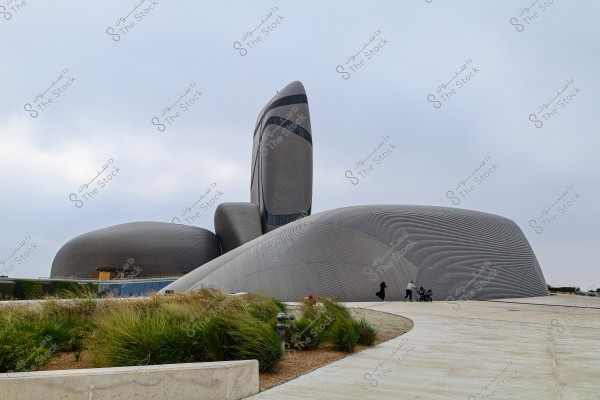 A modern building with a creative architectural design, featuring a unique and curved shape. The building is in muted gray and silver tones, set against a cloudy sky. In front of the building, there is a light-colored concrete pathway surrounded by green grass.