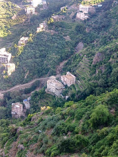 Aerial view of a mountainous village with white houses scattered among lush green hills filled with trees and plants. The area contains natural terraces used for agriculture. Narrow, winding dirt roads can be seen weaving between the farms and residences, indicating challenging terrain in the region.