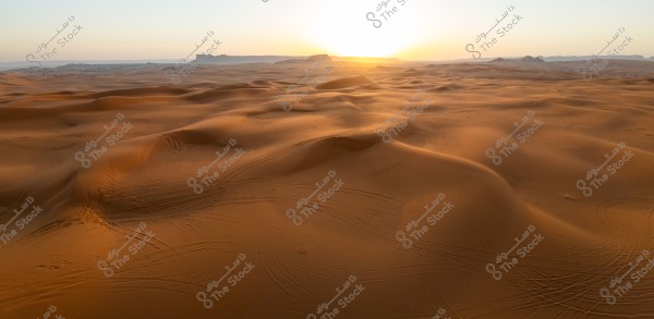 A natural landscape of a desert with multiple sand dunes stretching into the horizon, illuminated by the setting sun in the background. The horizon is clearly visible, with golden sandy colors blending into the gradient sky. Tire tracks can be seen on the sand, indicating the passage of vehicles or desert excursions.