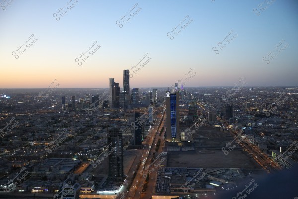 Aerial view of Riyadh in Saudi Arabia at sunset, featuring modern skyscrapers with sparkling lights. The sky gradually fills with an orange hue as light envelops the city. Buildings are scattered widely in the background.