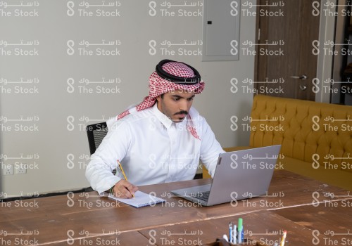 رجل سعودي يرتدي الزي السعودي التقليدي يجلس على طاولة الاجتماعات يستخدم الابتوب بينما يدون الملاحظات في الدفتر