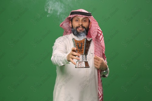 رجل سعودي يقف على خلفية خضراء يرتدي الثوب و الشماغ، يقوم بعمل حركات بيديه ، يبخر نفسه بالطيب و العود، احتفالات العيد