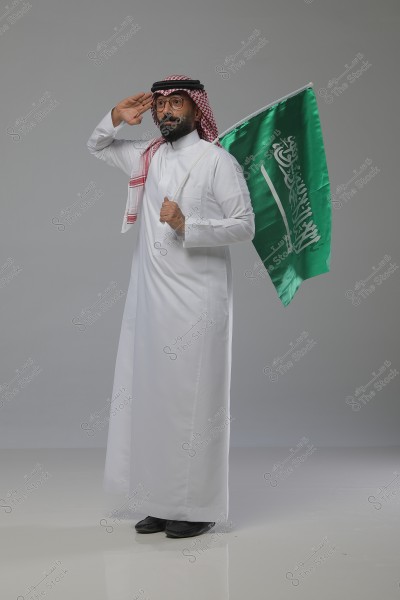 A portrait of a man wearing traditional Saudi white attire, with a red and white headscarf and a black agal. He is standing sideways and holding the green Saudi Arabian flag, performing a military salute. The background is light gray.