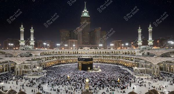 A nighttime view of the Grand Mosque in Mecca, showing the Kaaba surrounded by a large number of worshippers. The mosque is flanked by minarets and tall architectural buildings, with bright lights illuminating the entire area, while the night sky is adorned with shining stars.