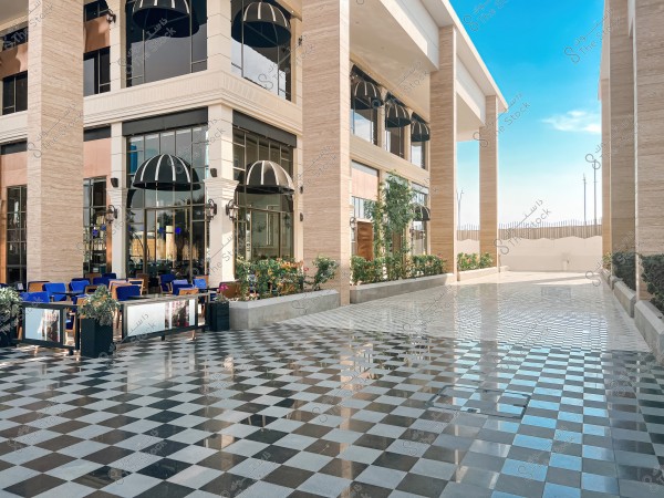 The image shows a modern outdoor courtyard featuring a black and white checkered tiled floor. On the right is a building with a glass facade and marble pillars, equipped with blue chairs and tables for outdoor dining. Plants and flowers are planted along the side, and in the background is a clear blue sky with a wooden fence.