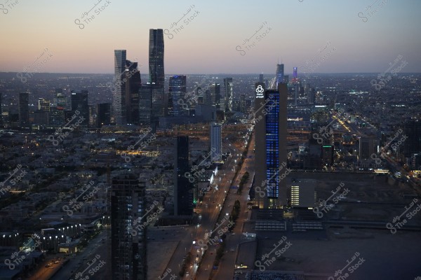 صورة لمدينة الرياض في المملكة العربية السعودية عند الغروب. تُظهر الصورة منظرًا جويًا يبرز الأبراج العالية والمباني الحديثة في وسط المدينة. الطريق مضاء بالأضواء والسيارات المتحركة, وتظهر مباني المدينة متراصة بتصميماتها العمرانية الحديثة.