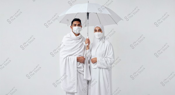 A photo of a man and woman wearing traditional white Ihram clothing; the man is in the Izar and Rida, and the woman is in full hijab. Both are wearing white face masks and holding an open white umbrella above their heads. The background is plain white.