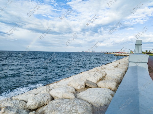 A scenic view overlooking the ocean with waves crashing against large white rocks forming part of the shoreline. In the distance, a port is visible with cranes and small buildings. The sky is partly cloudy with some sunshine, and a faint rainbow can be seen in the sky.