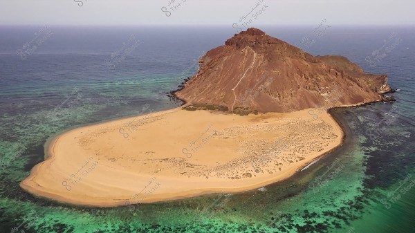 An aerial view of a small island situated in the blue sea. The island features soft golden sands and is surrounded by clear blue waters with visible coral reefs beneath the surface. The central part of the island is a brown rocky hill that adds distinct geographical features to the natural landscape.
