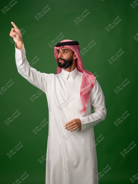 Portrait of a man wearing a white thobe and a red-checkered ghutra with an agal, standing against a green background. The man is pointing upwards with his finger. The outfit reflects traditional Saudi clothing.