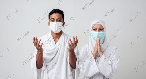 A photo of a man and a woman wearing traditional Ihram clothing. The man on the left is in a white robe, and the woman on the right is in a white hijab. Both are wearing medical masks. The man has his hands raised in prayer, while the woman has her hands together in a gesture of prayer.