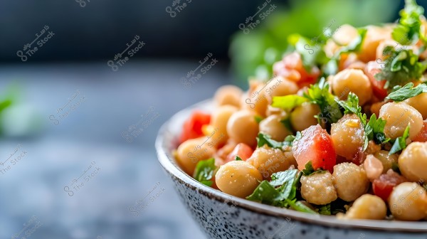 A close-up shot of a bowl containing a chickpea salad with chickpeas, diced tomatoes, and fresh parsley leaves. The background is blurred, highlighting the salad ingredients in the foreground.