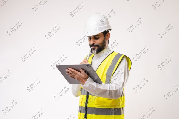 Image of a man wearing a white thobe, a yellow reflective vest, and a white hard hat, standing and holding a tablet in his hand, appearing to write notes. The man has a beard and is dressed in attire suggesting work in construction or engineering. The background is plain white, focusing on the man and the tablet he is using.