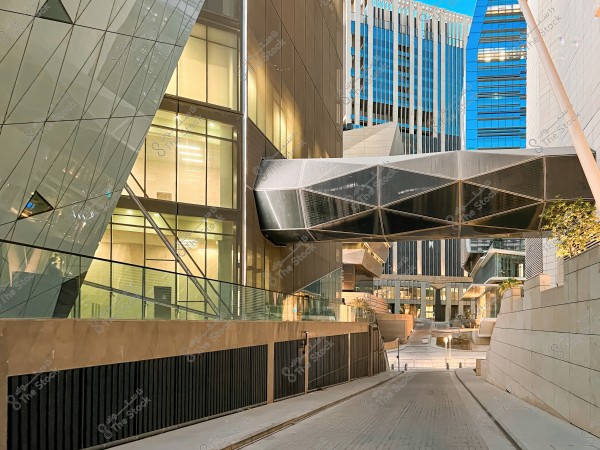 An image of a modern urban alley surrounded by glass buildings and contemporary architecture. A closed bridge with an angular design spans over the narrow paved road. The buildings reflect light and display multi-story windows with modern geometric details. The atmosphere suggests a sophisticated and advanced cityscape.