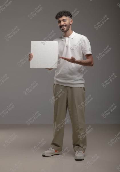 Portrait of a young man standing and smiling, wearing a white polo shirt and beige trousers, holding a square white board in his right hand. The background is gray, and the setting appears to be a studio.