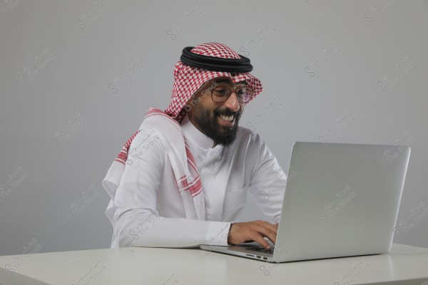 A portrait of a man sitting at a table working on a laptop. He is wearing a white thobe and a red and white ghutra with a black agal, indicating traditional Saudi Arabian attire. The man appears to be smiling and focused.
