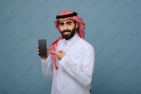 بورتريه شاب سعودي يرتدي الزي السعودي يشير إلى هاتفه ويتفاعل معه، وتظهر على ملامحه الدهشة ، على خلفية سماويه