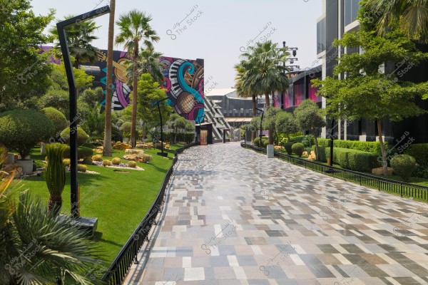 A decorative walkway with a colorful tiled ground passes through a green garden filled with various trees and plants. The surrounding walls feature large art murals with bright colors, depicting intertwined designs of musical elements such as strings, piano keys, and microphones. The scene is bright and appealing, suggesting a modern urban ambiance.