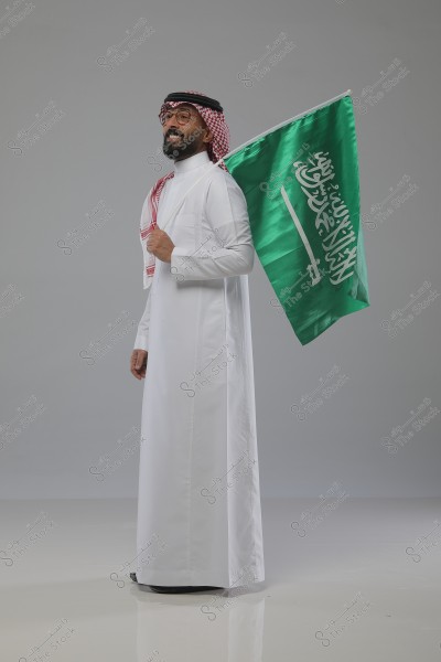 صورة لرجل يرتدي الزي السعودي التقليدي, الثوب الأبيض والغترة الحمراء والعقال الأسود, يحمل العلم السعودي. الخلفية رمادية بسيطة.