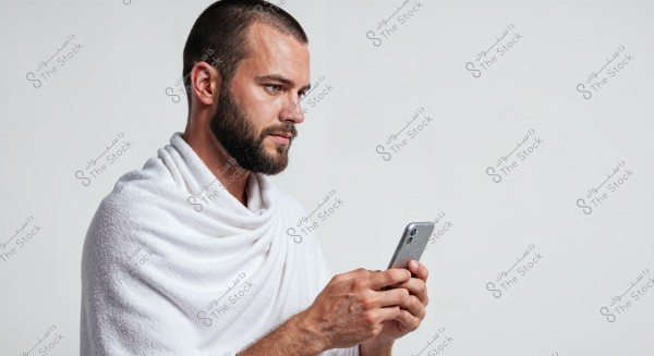 A portrait of a bearded man wearing a white towel, as if he has just taken a shower, holding a smartphone. The background is simple with a light gray color.