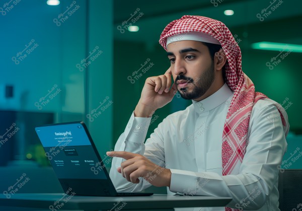 An image of a man wearing a white thobe and a red checkered ghutra, seated at a desk in a dimly lit conference room with green lighting. He appears focused on a laptop in front of him, with his finger pointing at the screen.