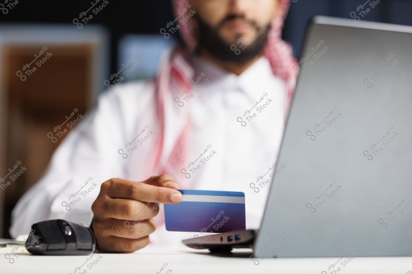 An image of a man wearing a white kandura and red checkered ghutra seated in front of a laptop, holding a blue credit card. He appears to be making an online purchase. A computer mouse is on the table beside him.