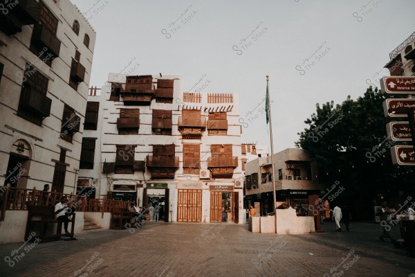 صورة لساحة مفتوحة في حي تاريخي, تُظهر مبانٍ تقليدية مكونة من طوابق متعددة مع نوافذ خشبية مزخرفة. يوجد علم بلون أخضر مرفوع على سارية طويلة. على اليسار, مجموعة من الأشخاص يجلسون على مقاعد تُطلّ على الساحة. تظهر أيضًا لافتات طريق توجيهية مكتوبة بالعربية والإنجليزية. الجو يبدو مشمسًا.
