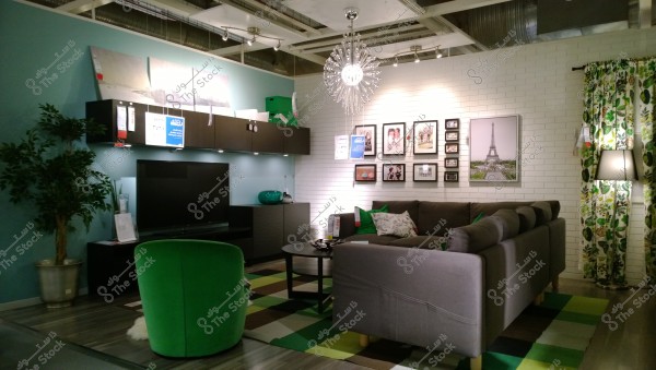 The image shows a modern living room featuring a large gray sofa adorned with green and white pillows. There is a round coffee table in front of the sofa with a multicolored rug underneath. The left wall is painted blue and has several art pieces hanging. There is a black TV unit with a large TV screen. In the left corner, there is a potted plant in a metallic pot. The right wall is covered with white brick and decorated with a collection of framed photos, including one of the Eiffel Tower. The curtains have a green botanical design, and a contemporary chandelier illuminates the room.