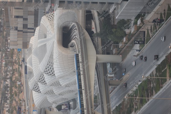 صورة لمحطة مترو حديثة بتصميم معماري فريد في مدينة حضرية. يغطي السقف ألواح بيضاء منحنية تتشابك بشكل فني. يمكن رؤية شوارع المدينة بجانب المحطة, مع سيارات تسير على الطرق المحيطة. تظهر المباني العالية المحيطة بالنباتات والأشجار, مما يضفي طابعًا حضريًا حديثًا.