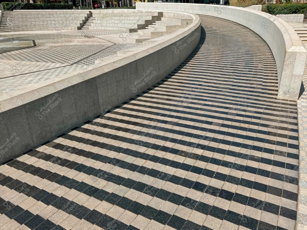 An open plaza featuring circular stone steps leading to a symmetrical seating area with alternating dark gray and light gray tile flooring. Green hedges are visible in the background.