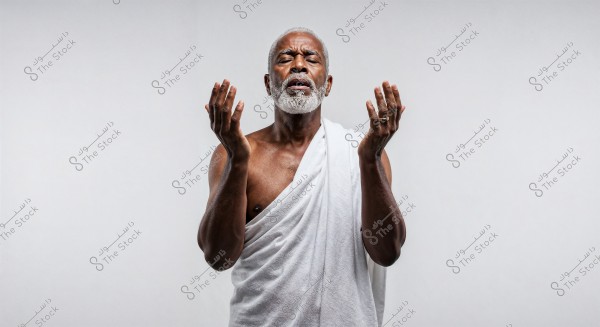 Image of an elderly dark-skinned man with a white beard, wearing a white Ihram garment, appearing in a state of meditation or prayer. The man has his eyes closed and his hands raised in front of his chest in a praying position. The background is simple and white.