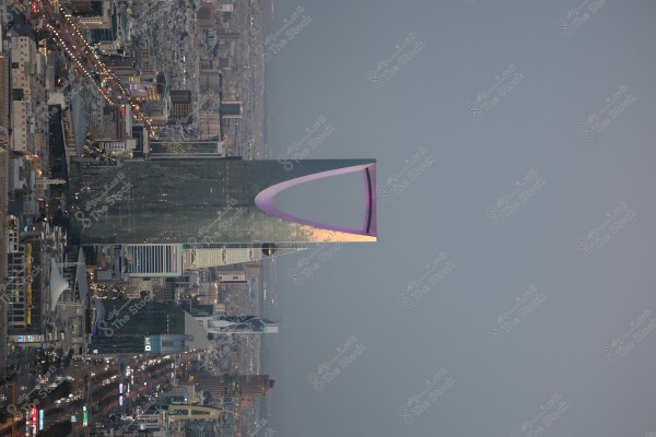 صورة ليلية لمدينة الرياض في المملكة العربية السعودية, وتظهر فيها أبراج ومعالم بارزة من ضمنها برج المملكة الشهير بإضاءته الأرجوانية المميزة على شكل قوس في الجزء العلوي. تنتشر الأضواء عبر المدينة, مما يبرز الشوارع والمباني المختلفة.