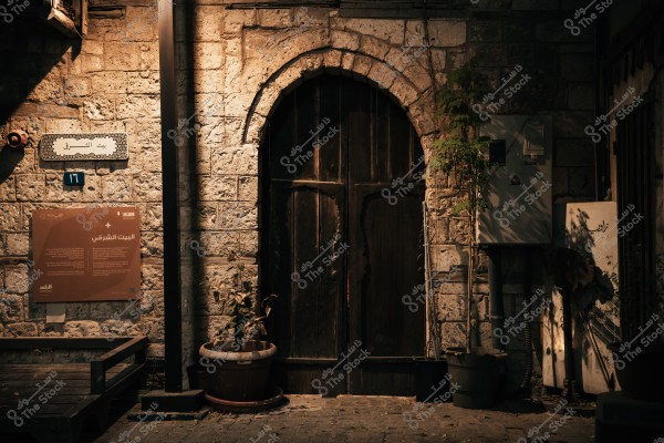 An image depicting the facade of a traditional building featuring an arched wooden door surrounded by stone walls. A dark-colored sign is displayed on the wall, providing information about the "Eastern House" in Arabic. Pots with small plants are placed beside the door, and there is a rectangular metal panel attached to the wall. The scene is illuminated with warm lighting, highlighting the architectural details.