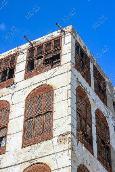 An image of an old building with traditional architectural style showcasing a side facade at the corner of the structure. The building features large, rectangular windows with brown wooden shutters. The exterior walls are slightly worn, showing signs of age, with a clear blue sky in the background.