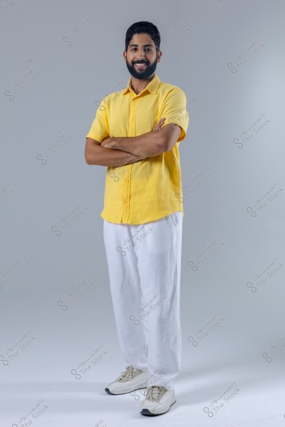 A portrait of a man standing with a smile and his arms crossed, wearing a yellow shirt, white trousers, and white sneakers, against a soft gray background. The simple, casual style may be fashionable in countries like Saudi Arabia.