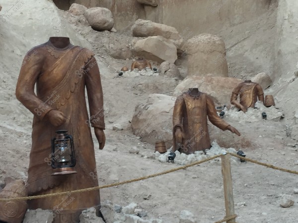 The image shows a group of headless wooden statues in a desert-like area. The statues depict figures in traditional clothing holding lanterns and other items, partially buried in the ground amid rocks and sand. The surrounding rocky edge creates an archaeological appearance.