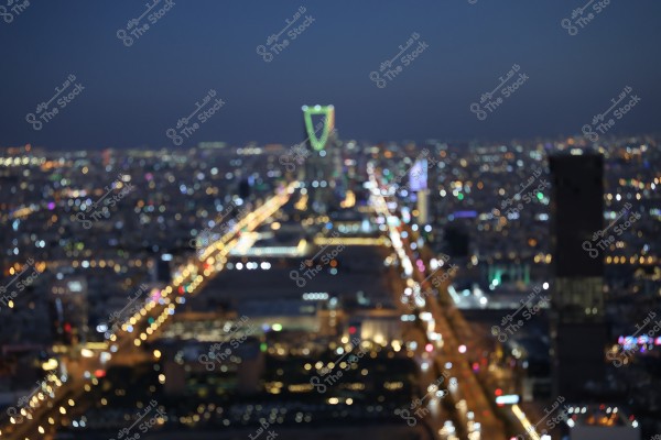 A nighttime view of a brightly lit city is depicted in the image, with a blurred effect making details hard to distinguish. The Kingdom Tower in Riyadh, with illuminated lights, is centered in the image, surrounded by the twinkling city lights in the background.