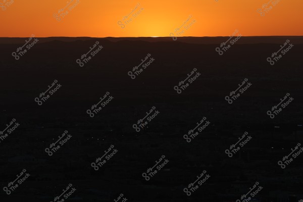 Sunset view over the horizon, with the sun disappearing behind distant hills, creating dark silhouettes in the foreground. The sky is filled with a warm, glowing orange hue, imparting a feeling of tranquility and natural beauty.