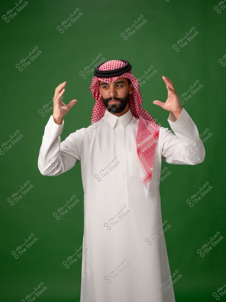 Image of a man wearing traditional Saudi white attire with a red headscarf and a black agal, standing against a green background. The man appears to be gesturing with his hands, his face showing focus.