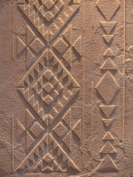The image shows decorative art carved into a clay wall with a brown color. The geometric design features triangular and diamond shapes arranged in a repetitive pattern, highlighting the fine details of the wall\'s texture. The style appears to be inspired by traditional architecture in the Middle East or North Africa.