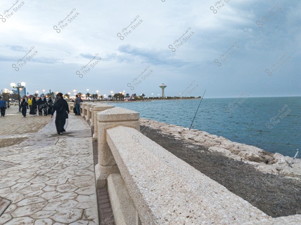 An evening view of a seaside promenade with a group of people wearing black abayas walking along the pavement next to the sea. On the right side, there is the sea water, and a tall structure is visible in the distance, accompanied by palm trees and lights in the background. The weather appears cloudy.