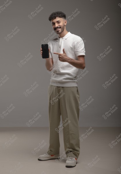 A photo of a man standing in a studio with a gray background, wearing a white polo shirt and beige trousers. He is smiling, holding a black smartphone, and pointing at it with his finger. He is wearing white sneakers.