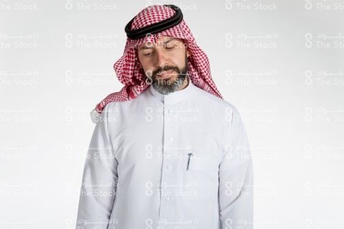 بورتريه رجل سعودي يقف على خلفية بيضاء، يرتدي الثوب، ينظر بعينيه نحو الاسفل، تعبير تفكير عميق