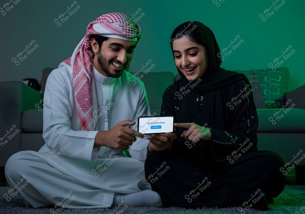 A man and a woman are sitting on the floor in a room next to a sofa. The man is wearing a white traditional thobe and a ghutra, while the woman is wearing a black abaya and hijab. Both are looking at a smartphone they are holding, with the phrase \"Confirm Purchase\" displayed on the phone\'s screen. They appear to be happy and cheerful.
