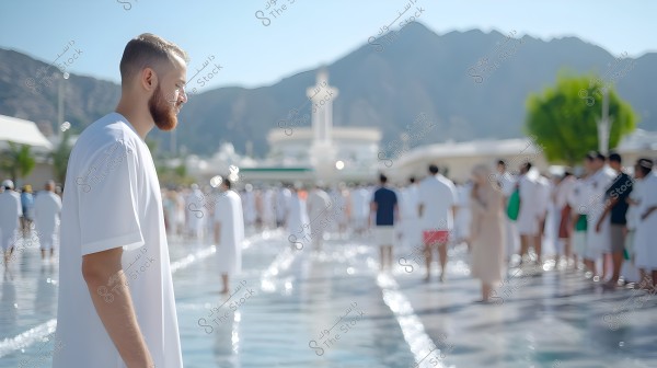 The image shows a man wearing a white thobe, possibly Saudi or Gulf origin, walking in a large water-covered plaza. In the background, a crowd of people is gathered, dressed in similar attire, suggesting a religious or celebratory atmosphere. Mountains and architectural landmarks can be seen in the distance under a clear blue sky.