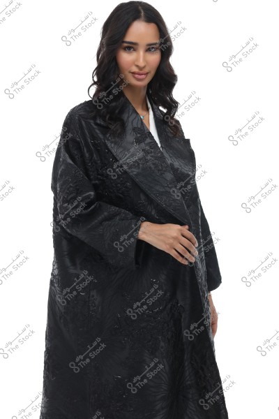 An image of a woman wearing a black abaya with elegant designs. The abaya appears traditional and luxurious, typical of women\'s wear in Gulf or Arab countries. The woman is wearing a simple necklace, and her long, wavy hair is black. She has a slight smile against a white background.
