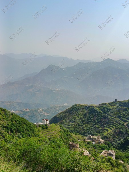 A scenic view of green terraced mountains under a clear blue sky. The hills are covered with varying shades of green foliage, and traditional buildings are scattered in some areas. The horizon features multiple layers of mountains extending into the distance.