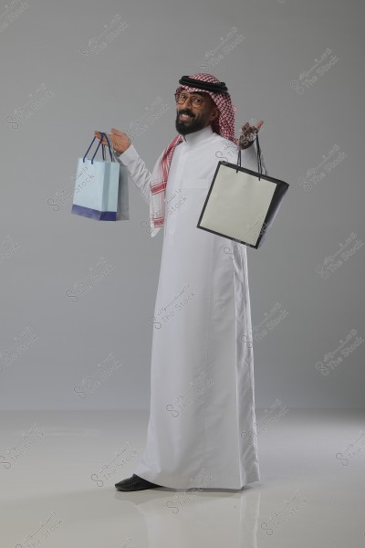 A portrait of a man wearing a white thobe and a red-checkered ghutra with a black agal. He looks happy as he smiles and holds shopping bags of different colors. He stands against a grey background.
