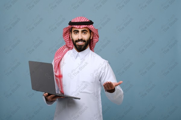 بورتريه شاب سعودي يرتدي الزي السعودي  يحمل كمبيوتر محمول ، يقف على خلفية سماوية